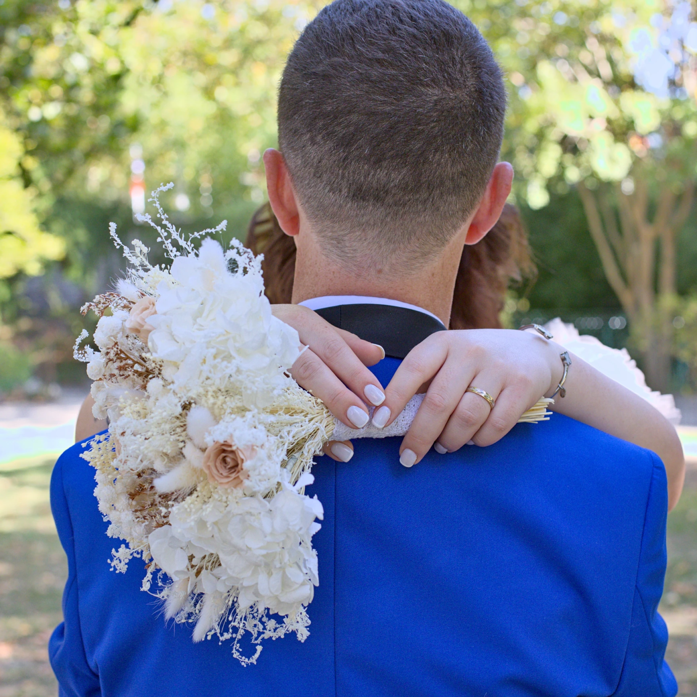 Moment intime La mariée enlace le cou du marié avec le bouquet à la main. La photo montre le marié de dos et met en valeur les détails du bouquet et les mains.