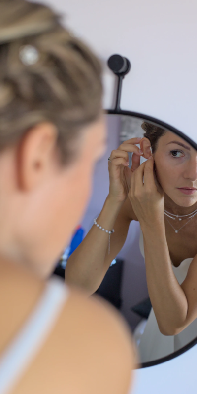 Les derniers ajustements La mariée ajuste ses boucles d'oreilles de mariage face au miroir. Une photographie qui à capturé un moment original des préparatifs.