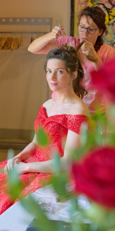 Rose rouge et robe rouge Photo d'une mariée vétue d'une robe rouge prise à travers des roses. On voit la mariée en train de se faire coiffer par une coiffeuse durant les préparatifs à Grenoble.